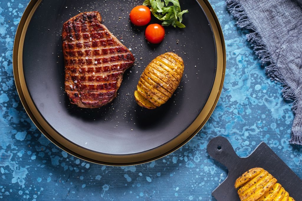 Biftek sa hasselback krompirom i čeri paradajzom na tamnom tanjiru, pogled odozgo